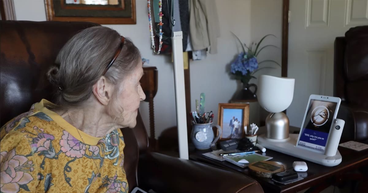 Older woman with gray hair and a yellow dress looking at a photograph on a dresser.