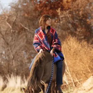Dr. Em Loerzel rides a horse at sunset. She is wearing a heavy red and blue shawl.