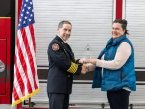 South County Fire Chief Shaughn Maxwell and Jenn Stuber pose with her award, both holding the award between them, and smiling for the camera. Stuber is wearing a puffy blue vest and Maxwell is in uniform.