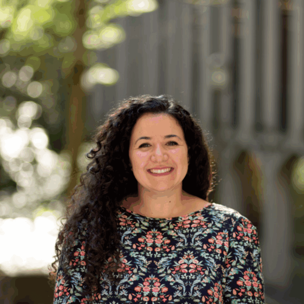 A young woman smiles, wearing a red and blue patterned shirt. She's outside, in front of a university building on a sunny day.