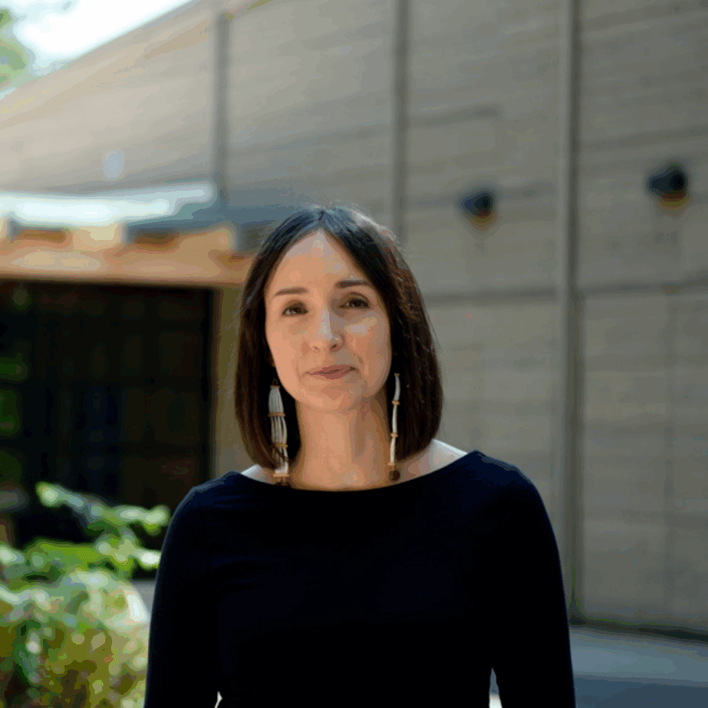 A young dark-haired woman wearing a black sweater looks into camera. She's in front of a building on a sunny day.