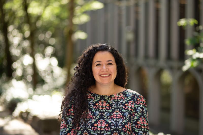 Sarah Porter smiles, wearing a multi-colored patterned shirt, while standing in a shaded area outside on a sunny day. A large university building is in the background.