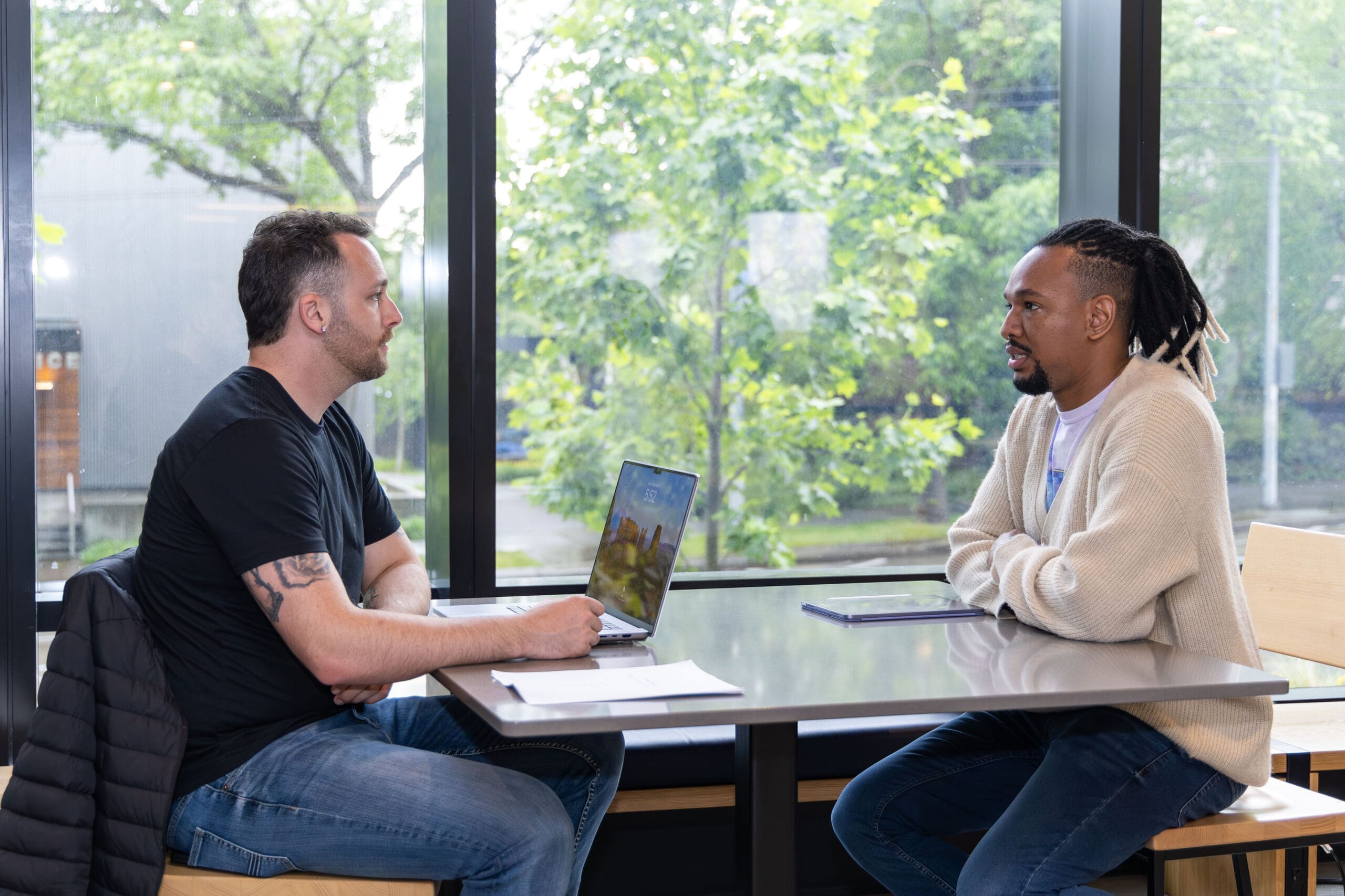 Two men sit at a table in a dormitory common area near a window on a sunny day. A laptop is open in front of one of them.