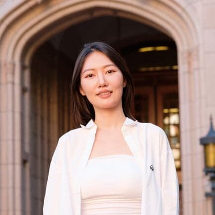 Dasol Kang, PhD Candidate for the UW School of Social Work, standing outside in front of a beige building, wearing a white jacket and white t-shirt.
